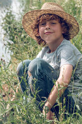 Delighted kid with curly hair and in straw hat sitting among green grass on channel coats and smiling while looking at camera in sunny summer day - ADSF47379