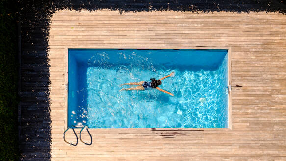 Drone view full length of unrecognizable woman with long wet dark hair floating in outdoor swimming pool on sunny day in tropical resort - ADSF47381