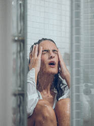 Upset female with dark hair in white wet shirt crying while sitting in shower under water stream with closed eyes and touching head - ADSF47382