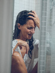 Dressed long haired lady crying in shower cabin while sitting under spraying water and touching forehead - ADSF47384