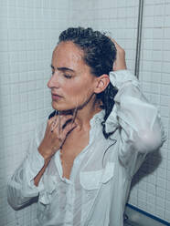 Thoughtful female in wet shirt standing with closed eyes under splashing water in shower cabin and touching head - ADSF47386