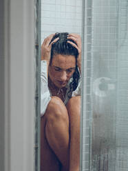 Exhausted female in white wet shirt sitting on floor of shower cabin and touching head with hands under pouring water - ADSF47387