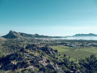 Picturesque drone view of Pollensa town located amidst green agricultural fields and mountains against blue sky on coast of sea in Mallorca, Spain - ADSF47388