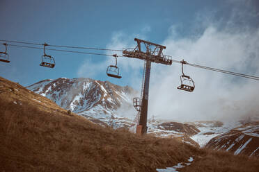 Ropeway with seats for transporting tourists going under grassy hills surrounded by snowy mountains covered with steam - ADSF47389