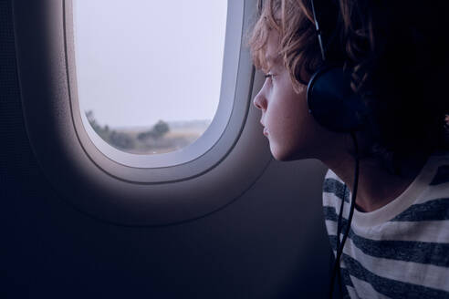 Kid listening to music in headphones and looking out window of dark plane during flight in evening - ADSF47390
