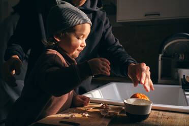Side view of funny little toddler in casual clothes and hat peeling fresh ripe oranges standing at table with crop anonymous father in kitchen - ADSF47392
