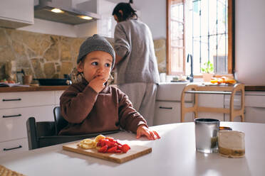 Adorable little child sitting on chair at table and eating delicious cut banana and strawberry against mother cooking food in kitchen - ADSF47393