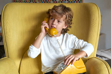 Content preteen child in casual outfit having conversation on retro telephone while sitting in comfortable armchair in light living room at home and looking away - ADSF47395