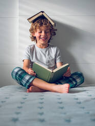 Full body of smiling little boy in nightwear sitting on bed with crossed legs and reading interesting story with book covering head - ADSF47399