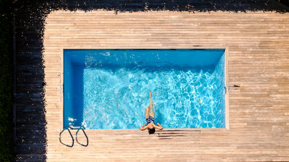 Aerial top view of unrecognizable female tourist with wet dark hair in swimwear leaning on wooden deck of outdoor pool and splashing water with legs during summer holidays - ADSF47401