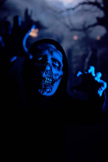 Awful undead monster stretching out arms and limping towards camera under blue neon light at night on dark cemetery - ADSF47404