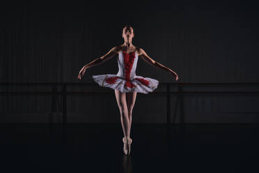 Full body of slim woman ballet dancer in tutu skirt performing tiptoes movements while dancing in dark studio - ADSF47405