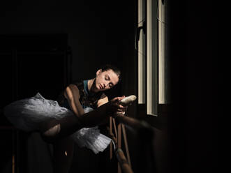 Side view of young female ballerina in tutu stretching leg on barre while training in dark studio - ADSF47407