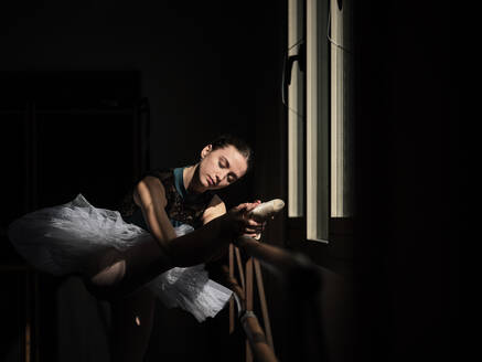 Side view of young female ballerina in tutu stretching leg on barre while training in dark studio - ADSF47407