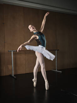Ganzkörperansicht von Seitenansicht anmutige Ballerina in weißem Tutu stehend mit erhobenen Händen auf Zehenspitzen in Spitzenschuhen während einer Probe im Studio - ADSF47409