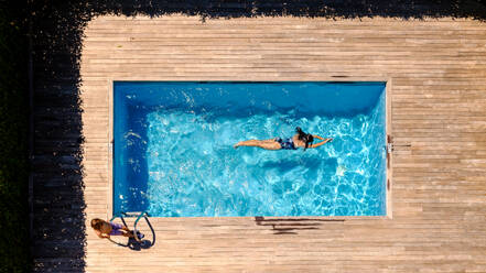 Drone view of anonymous little boy standing on wooden platform near young mother swimming in pool on sunny day during summer vacation - ADSF47411