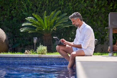 Side view of adult male freelancer sitting on edge of swimming pool and working on tablet with keyboard dock - ADSF47416