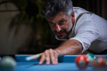 Bearded adult male in white shirt leaning on blue pool table and preparing to strike ball with cue while playing snooker in dark living room at home - ADSF47419