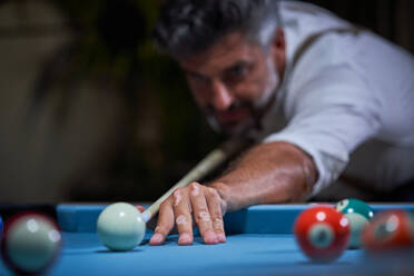 Crop serious man in white shirt standing at blue pool table and preparing to strike ball with cue while playing billiards in dark living room at home - ADSF47420