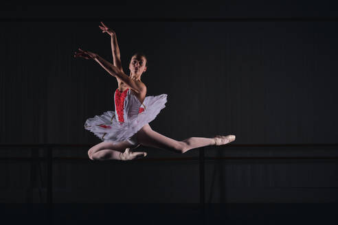 Ganzkörperansicht einer anmutigen Ballerina in Kostüm, Tutu und Spitzenschuhen, die mit erhobenen Händen tanzt, während sie eine Ballettrolle in einem dunklen Studio probt - ADSF47421