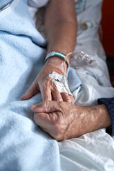 High angle of crop anonymous elderly man holding hand of sick woman with connected venous catheter lying on bed during rehabilitation treatment in hospital - ADSF47423