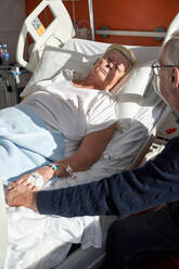 From above of crop senior man holding hand of unwell wife lying on medical couch with connected venous catheter during rehabilitation process in modern clinic - ADSF47425