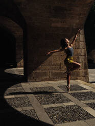 Full body side view of anonymous female dancer performing ballet dance movement on paved street with shadow and bright sunlight - ADSF47427