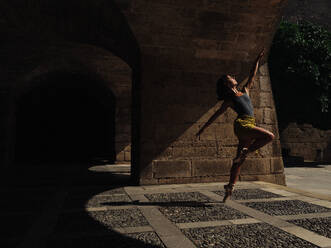 Full body side view of unrecognizable female performing ballet dance on paved street with shadow near shabby building in city - ADSF47428