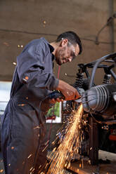 Side view of serious man in uniform with protective goggles standing at workbench and cutting metal of motorcycle with grinder while working in professional workshop on sunny day - ADSF47436