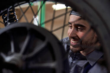 Low angle of positive bearded male mechanic in uniform and cap fixing problems in motorcycle wheel during workday in service - ADSF47441