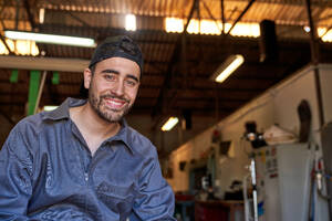 Professional male mechanic in workwear and cap smiling and looking at camera while sitting in garage on blurred background - ADSF47442