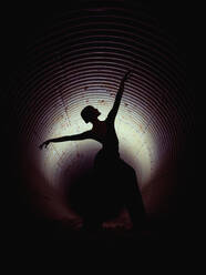 Silhouette of faceless woman dancing with raised arms in dark underground tunnel illuminated by light - ADSF47443