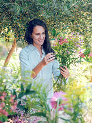 Glad brunette in white shirt creating bouquet of gentle flowers on sunny day in nature - ADSF47447