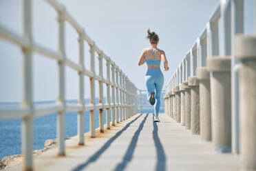 Back view of anonymous fit female runner in sports clothes jogging on fenced bridge over sea during training in sunlight - ADSF47457