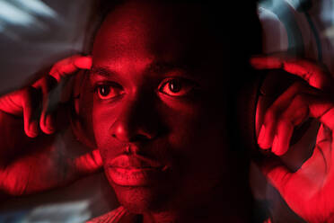 Serious African American male listening to music in wireless headphones in darkness with red illumination - ADSF47459