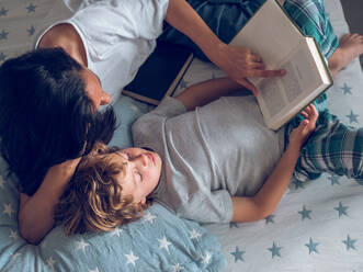 Top view of preteen boy and mom reading interesting book while lying on bed - ADSF47464