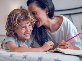 Happy cute preteen boy with curly hair smiling while drawing with cheerful mother - ADSF47465