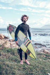 Full length of happy young bearded ethnic male surfer with curly hair in wetsuit smiling while holding surfboard on rocky coast near wavy ocean - ADSF47468