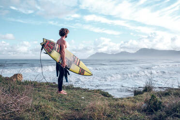 Side view of dreamy muscular shirtless male surfer with curly hair standing on grassy coast and with surfboard and admiring picturesque waving ocean against cloudy blue sky - ADSF47469