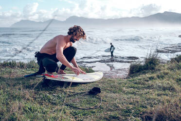 Side view of smiling barefoot and naked torso male surfer squatting and laying wax on surfboard on coast of wavy ocean - ADSF47470