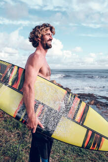 Side view of cheerful fit Hispanic guy with beard and naked torso smiling brightly while standing on grassy seashore with surfboard and looking away - ADSF47472