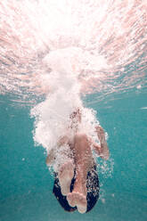 Underwater full body shot of unrecognizable barefoot kid wearing swimming shorts plunging in transparent clean blue water in pool - ADSF47476