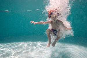 Full body of barefoot kid with closed eyes plunging under clean transparent water in pool while jumping against blue wall - ADSF47477