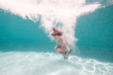 Side full body view of barefoot kid plunging in clean transparent blue pool water while jumping and embracing knees - ADSF47478