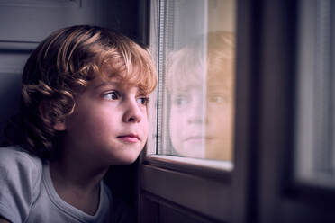 Adorable pensive little boy with curly blond hair leaning on window and looking away dreamily at home - ADSF47480