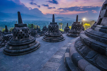 Amazing view of ancient largest Buddhist Borobudur Temple in Java Indonesia with bright focusing light during sundown time against orange cloudy sky - ADSF47494