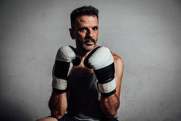 Portrait of strong adult male in sportswear looking at camera while sitting against gray wall with gloves hands near chin in daylight - ADSF47495