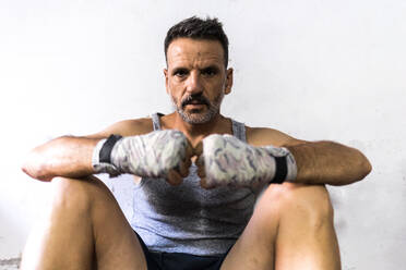 Serious adult male in sportswear looking at camera while sitting on floor leaning on white wall in room with wrapped fists together on knee in daylight - ADSF47496