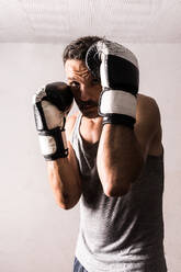 Confident adult male boxer looking at camera while standing near wall in room and keeping hand fists in boxing gloves near face in daylight - ADSF47498
