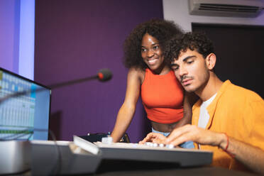 Male audio engineer and joyful female African American singer creating song with sound software while pressing buttons on mixing board in control room - ADSF47501